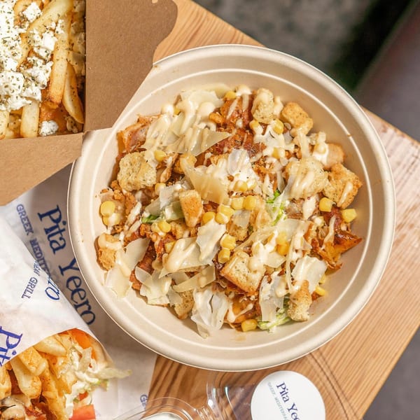 A Pita Yeero bowl with chicken, corn, croutons, cheese, and sauce, next to feta fries.