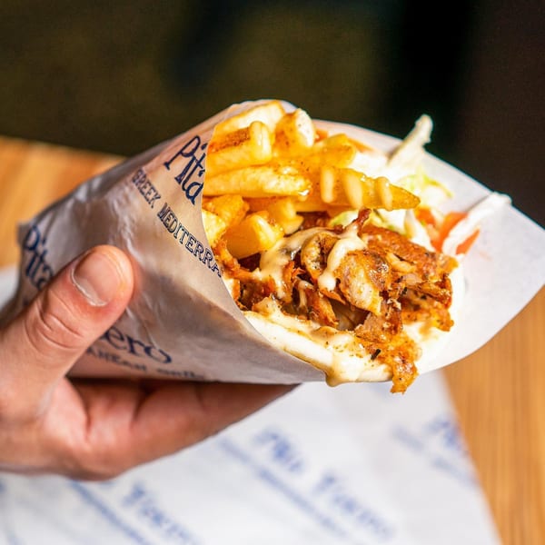 A hand holds a Pita Yeero, filled with meat, fries, and sauce, wrapped in branded paper.